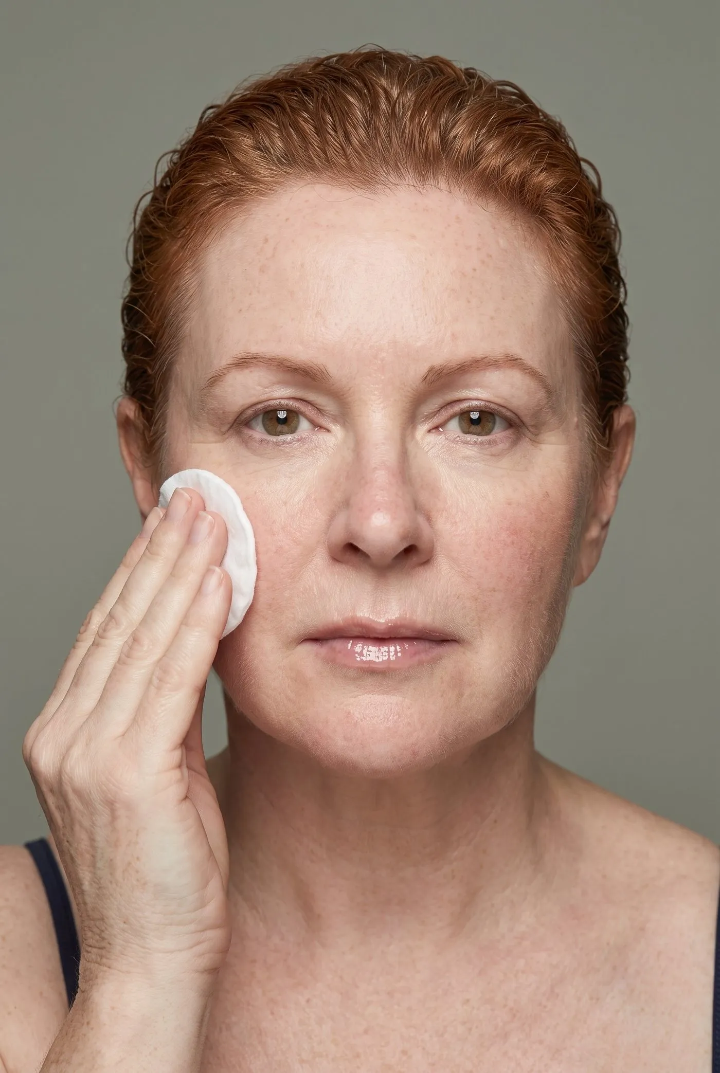 Studio photograph, a red-haired woman in her fifties, removing cleanser with a cotton pad, bare skin unretouched.