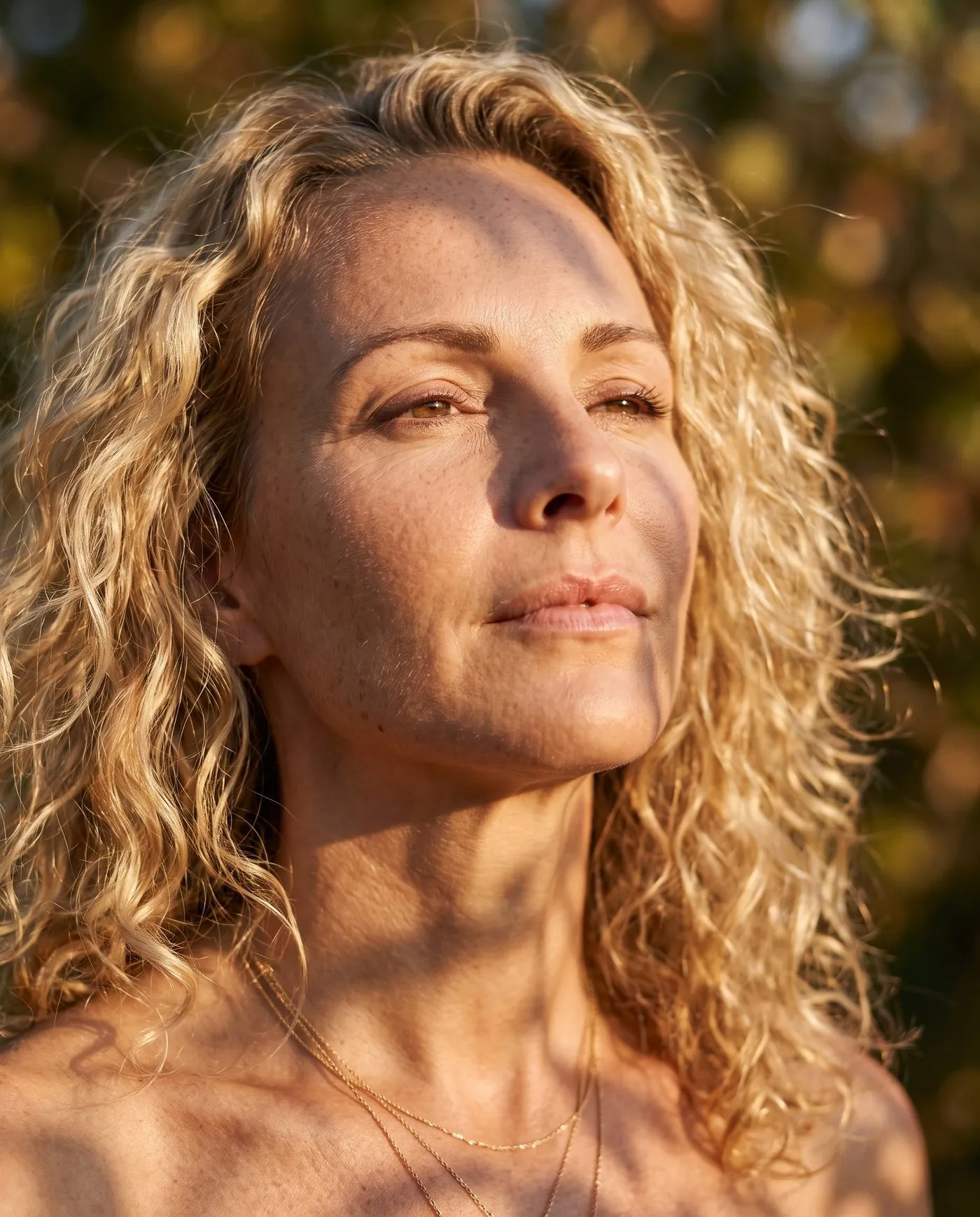Daylight portrait, a fair-haired woman in her forties, dappled leaf shadow falling across the face and neck, fine lines visible.