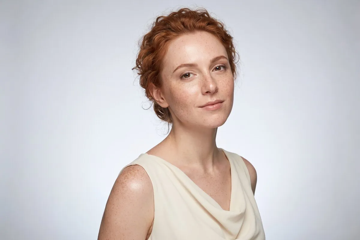 Composed studio portrait, a red-haired woman in her twenties, head and shoulders against a pale grey backdrop.