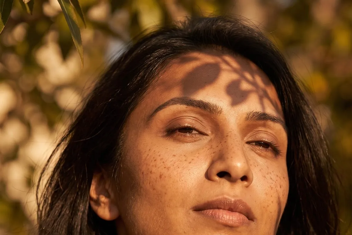 Daylight portrait, a dark-haired woman in her thirties, dappled leaf shadow falling across the face and neck, fine lines visible.