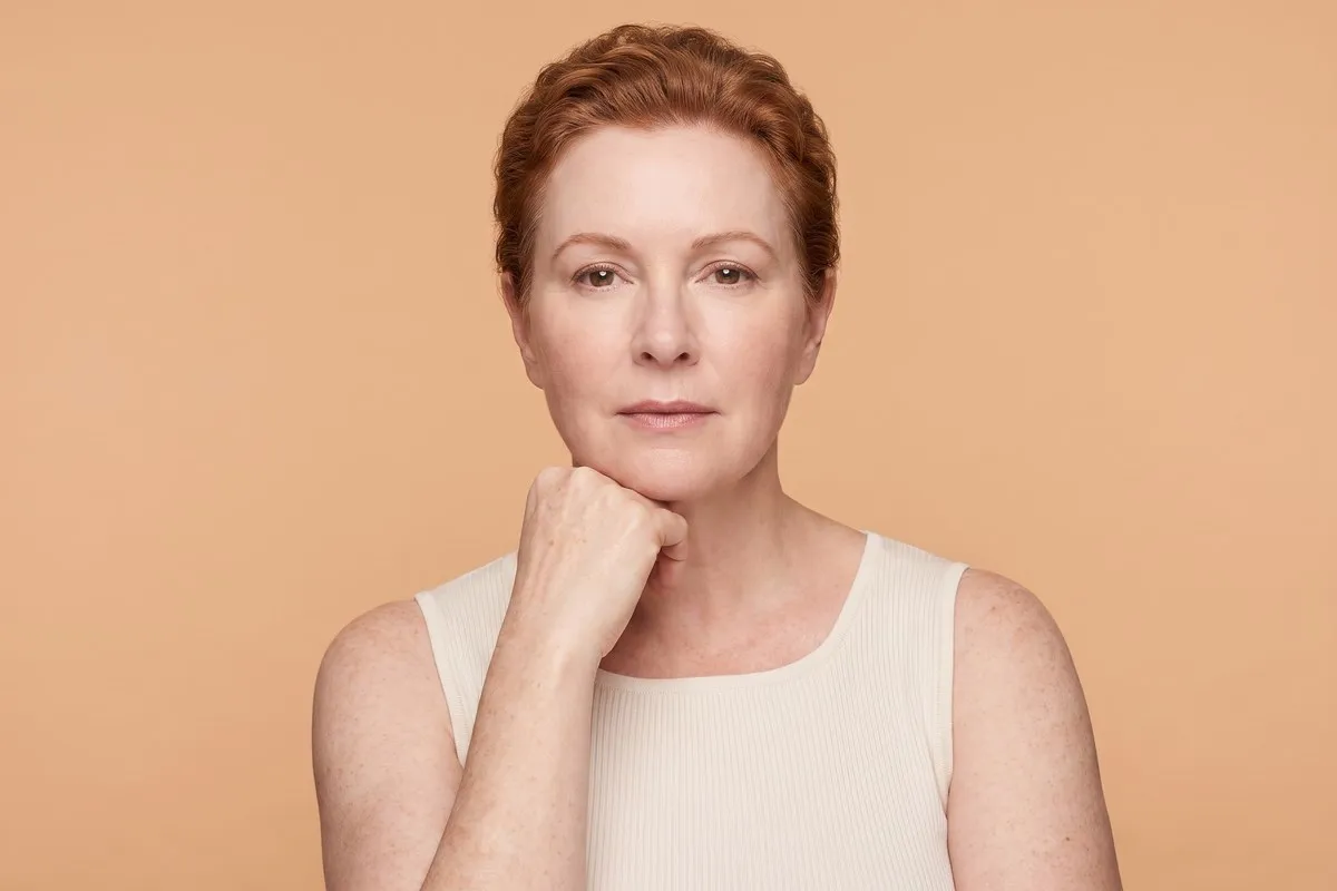 Studio portrait, a red-haired woman in her fifties, one knuckle resting beneath the jaw, against a warm sand backdrop.