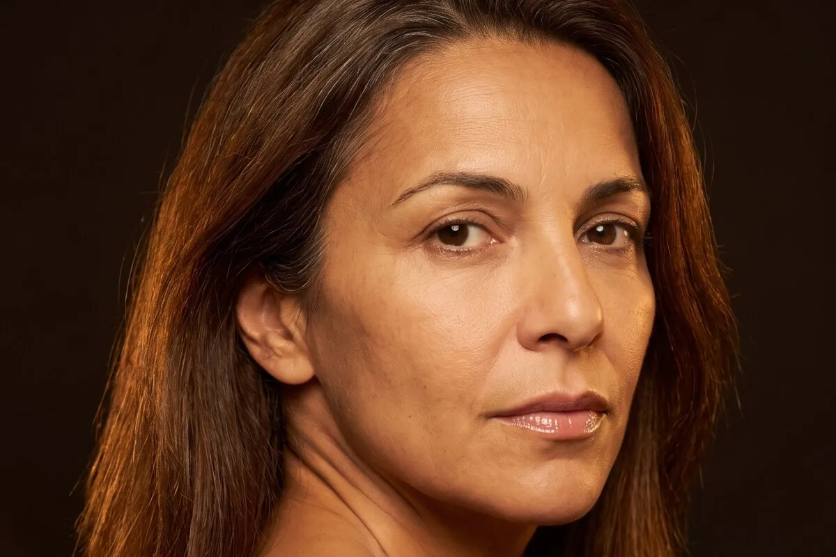 Low-key studio portrait, a dark-haired woman in her fifties, warm light falling across one shoulder.