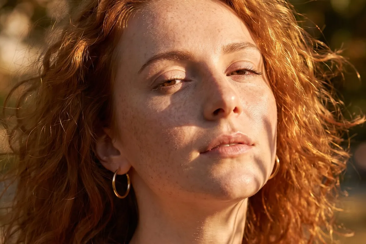 Daylight portrait, a red-haired woman in her twenties, dappled leaf shadow falling across the face and neck.