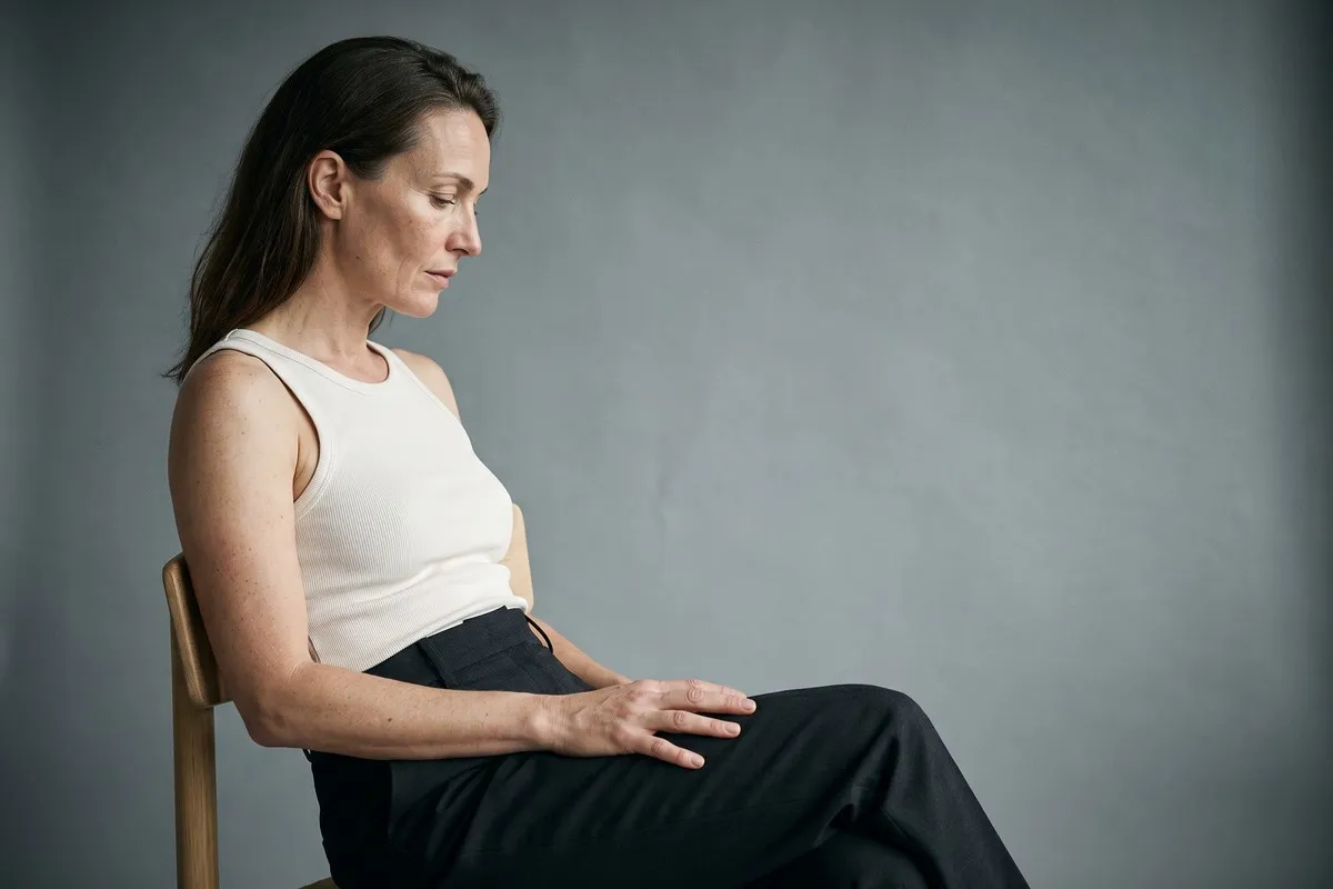 Studio photograph, a dark-haired woman in her forties, seated on a wooden chair in a plain vest, looking down, against a grey wall.
