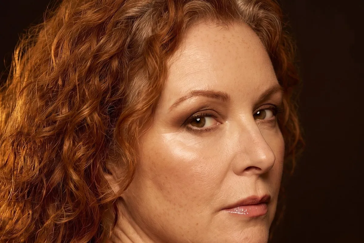 Low-key studio portrait, a red-haired woman in her fifties, warm light falling across one shoulder.