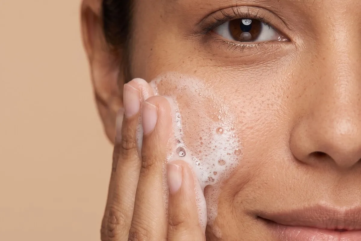Close studio photograph, a dark-haired woman in her twenties, cleansing foam on one cheek, bare skin unretouched.