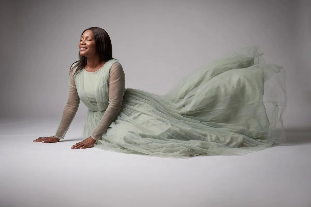 Studio photograph, a dark-haired woman in her fifties, seated on the floor in a long sage gown against a pale sweep.