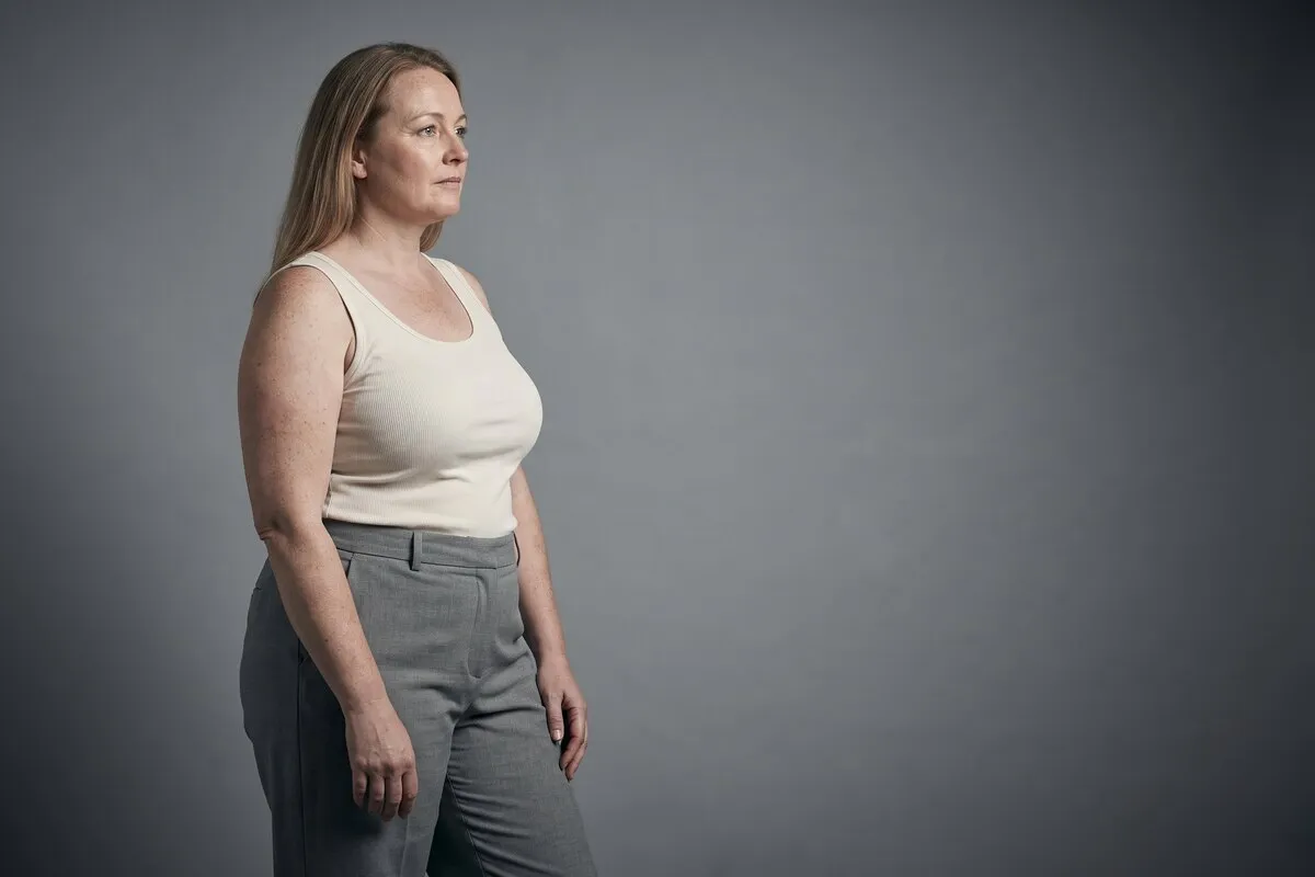Full-length studio photograph, a fair-haired woman in her forties, standing in plain clothing against a grey wall.