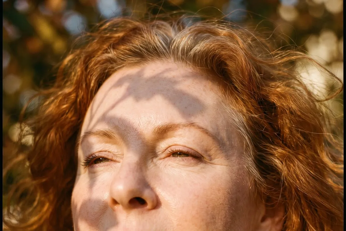 Daylight portrait, a red-haired woman in her fifties, dappled leaf shadow falling across the face and neck, fine lines visible.