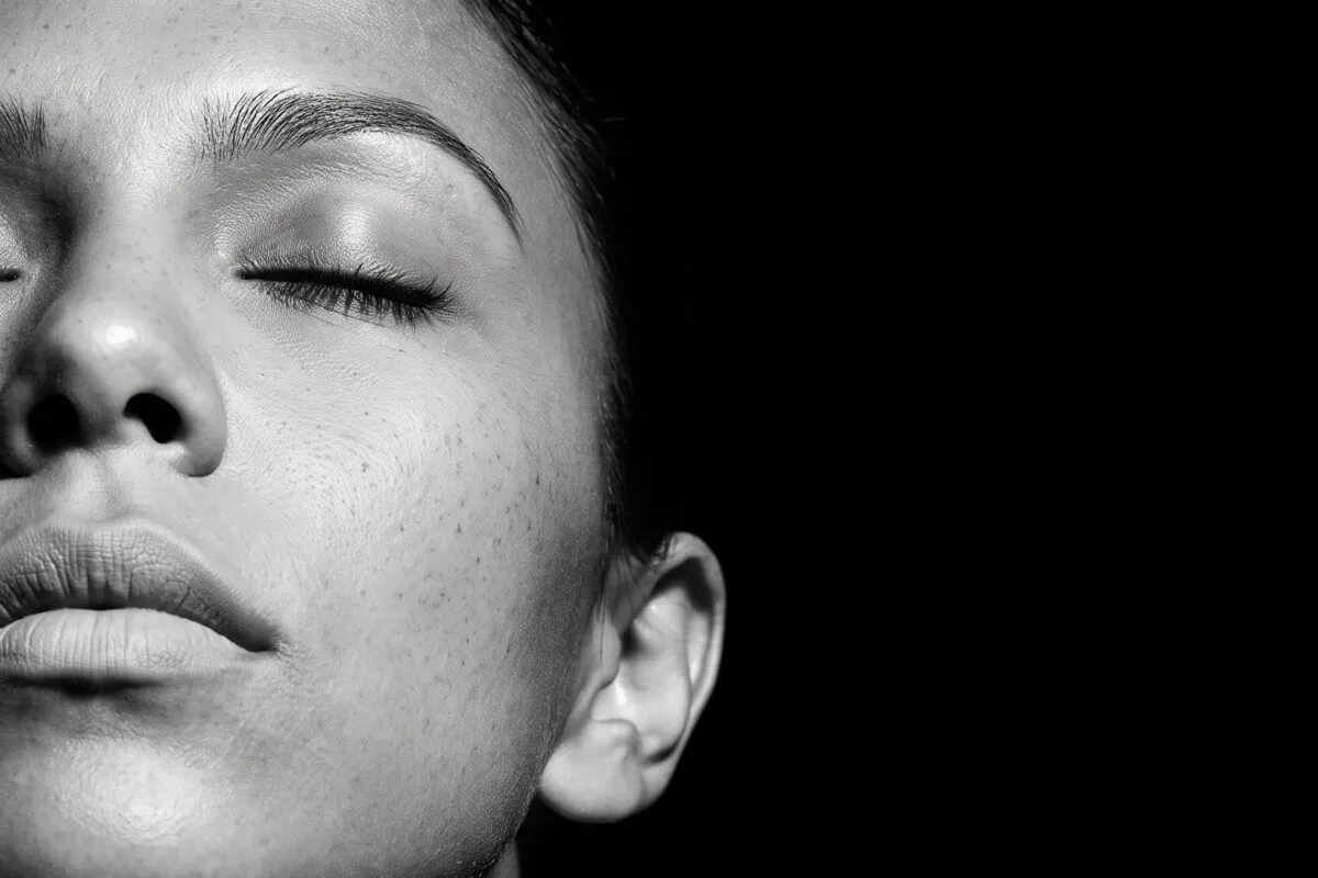 Black and white studio photograph, a dark-haired woman in her twenties, eyes closed against a dark ground, freckles and skin texture unretouched.