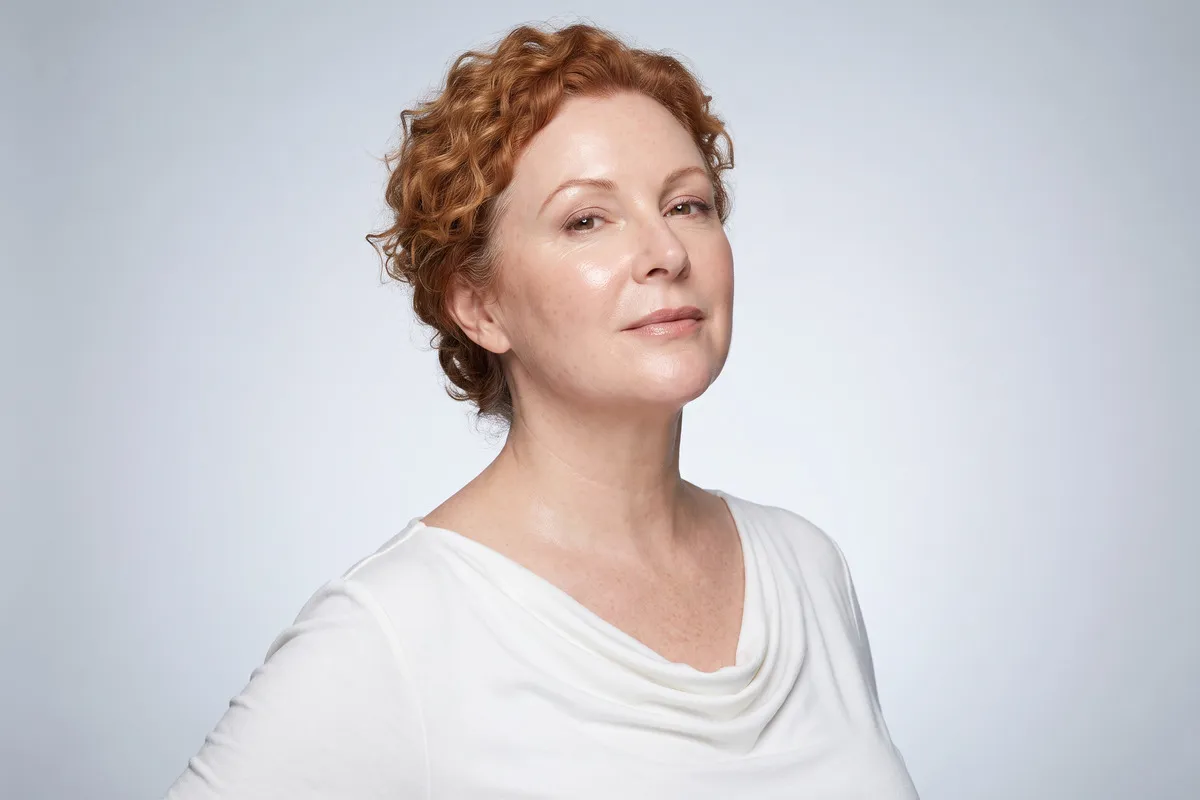 Composed studio portrait, a red-haired woman in her fifties, head and shoulders against a pale grey backdrop.