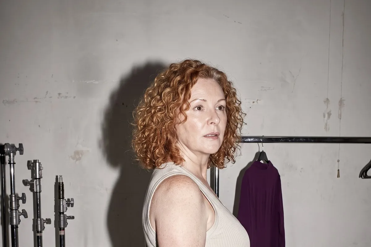 Backstage photograph, a red-haired woman in her fifties, standing beside a clothes rail and lighting stands against a plain wall.