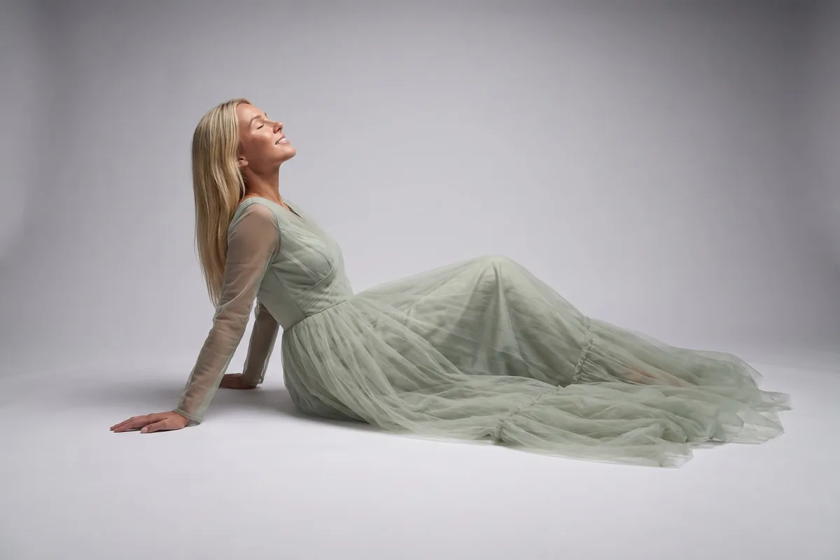 Studio photograph, a fair-haired woman in her twenties, seated on the floor in a long gown against a pale sweep.