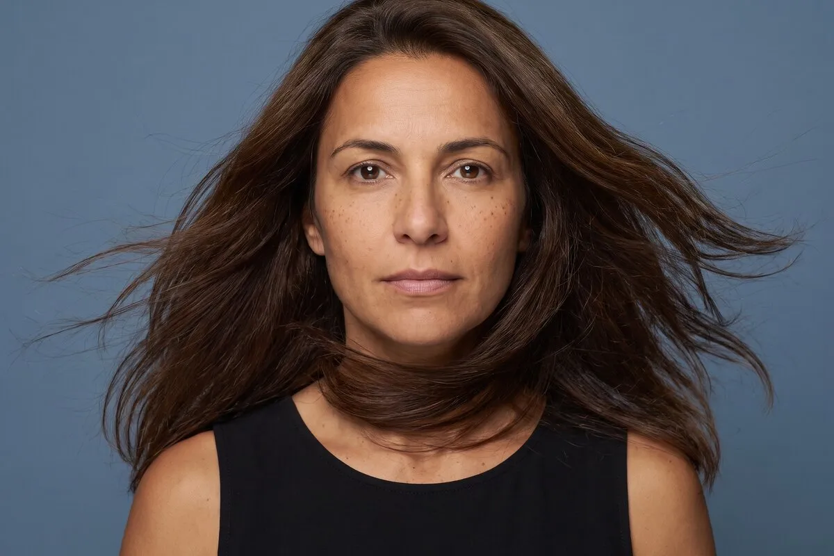 Studio portrait, a dark-haired woman in her fifties, hair caught mid-movement, against a blue-grey backdrop.