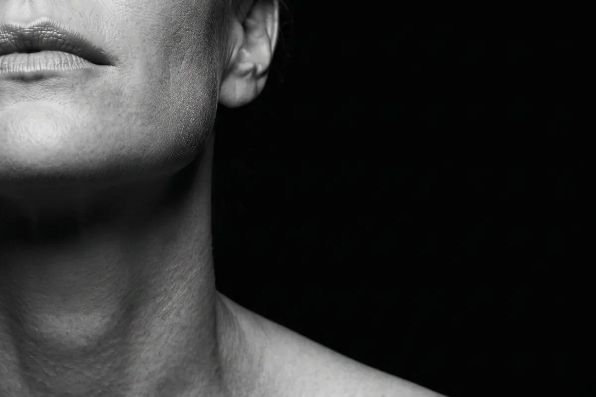Black and white studio photograph, a fair-haired woman in her forties, neck and collarbone against a dark ground.