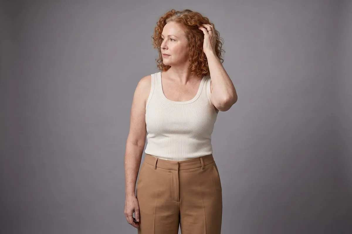 Full-length studio photograph, a red-haired woman in her fifties, standing in plain clothing against a grey wall.