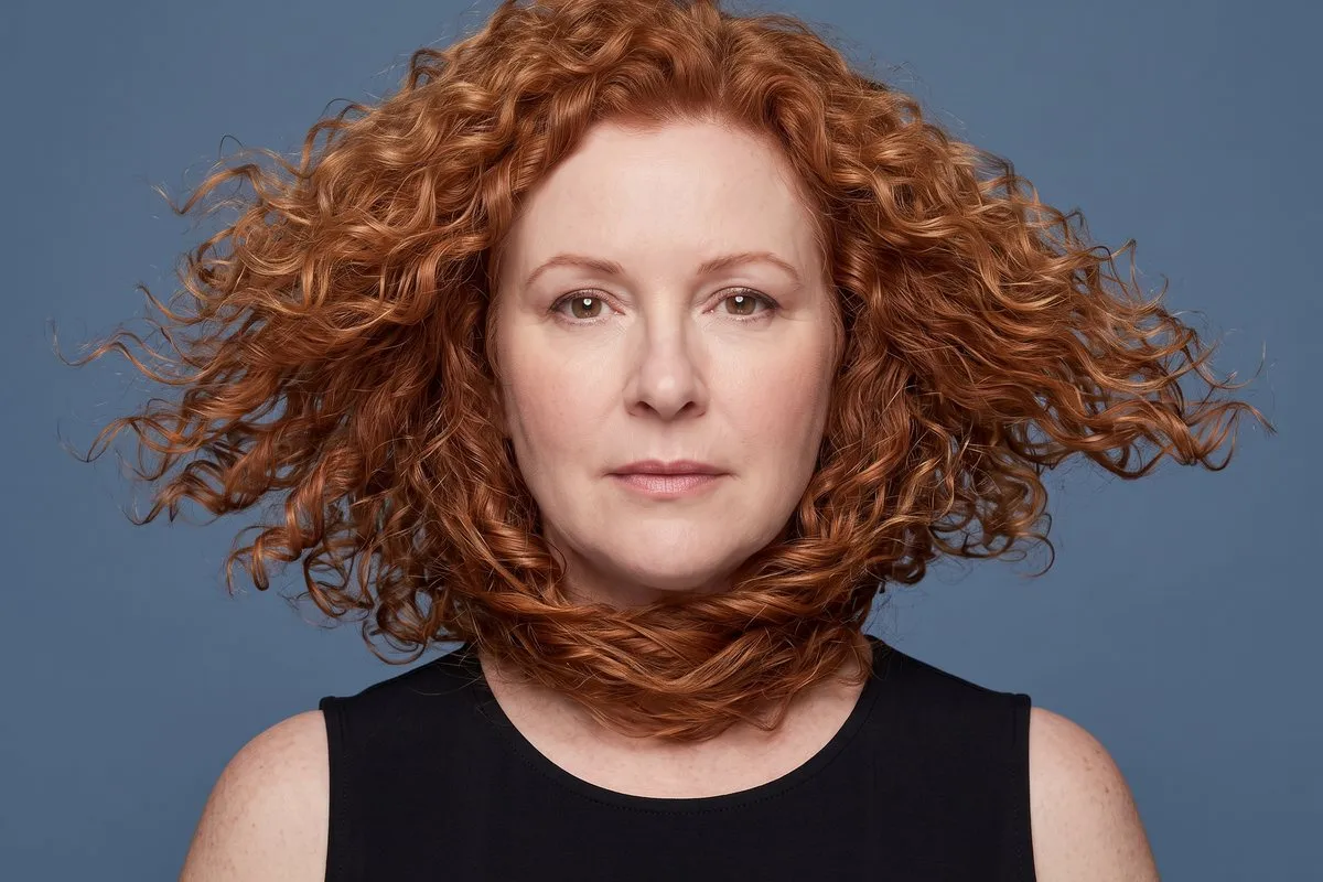 Studio portrait, a red-haired woman in her fifties, curls caught mid-movement, against a blue-grey backdrop.