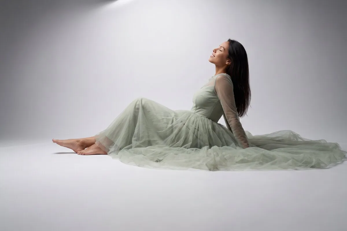Studio photograph, a dark-haired woman in her twenties, seated on the floor in a long gown against a pale sweep.
