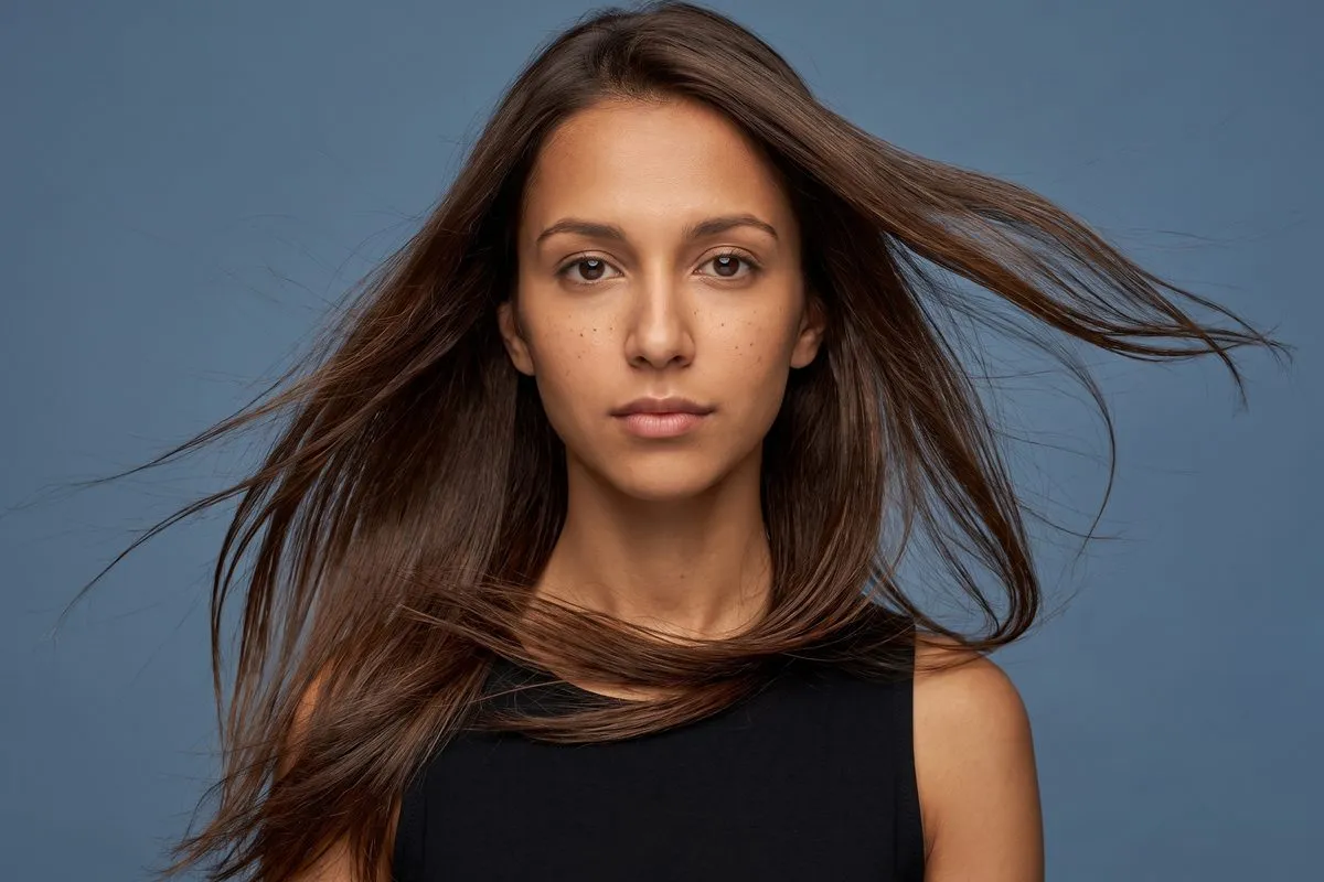 Studio portrait against a blue backdrop, a dark-haired woman in her twenties with her hair lifted by moving air.