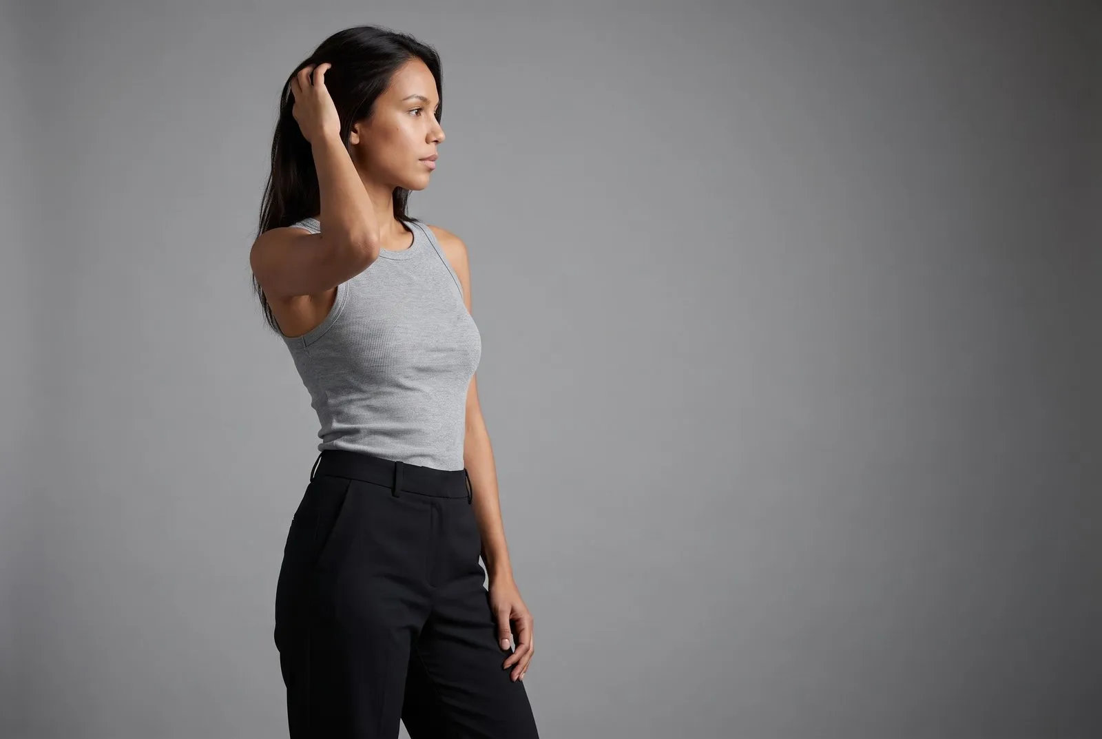 Full-length studio photograph, a dark-haired woman in her twenties standing in a grey vest and dark trousers against a plain grey wall.